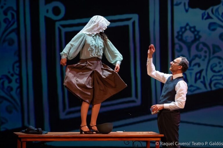 Una intérprete con pañuelo en la cabeza, de pie sobre una mesa, y un actor arrodillado que le tiende la mano. Foto: Raquel Cavero / Teatro Pérez Galdós