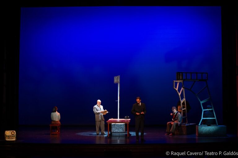 Grupo de intérpretes en penumbra azul alrededor de una mesa con un micrófono de pie. Foto: Raquel Cavero / Teatro Pérez Galdós