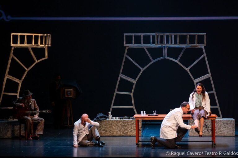 Un personaje arrodillado atiende a otro tendido en el suelo, junto a una mesa con objetos de laboratorio. Foto: Raquel Cavero / Teatro Pérez Galdós