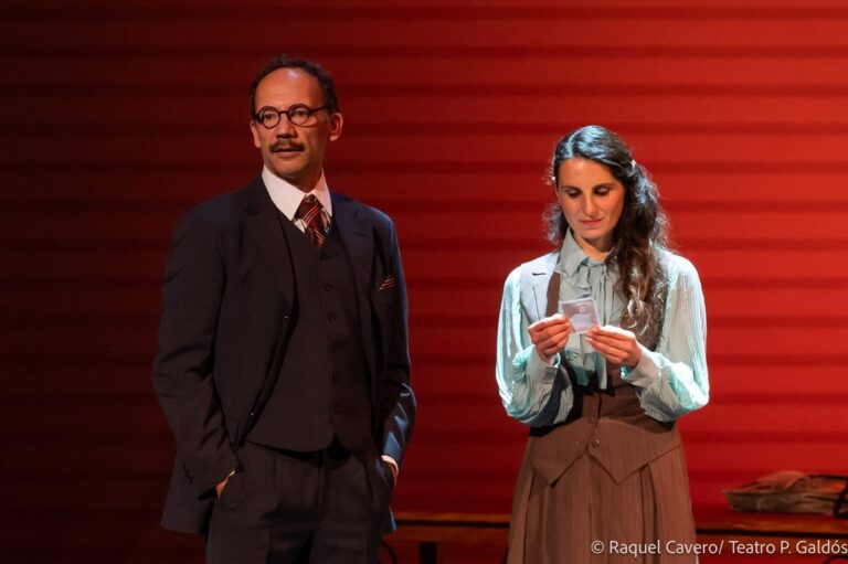 Dos intérpretes en primer plano sobre fondo rojo: el de traje oscuro y gafas, ella con blusa blanca. Foto: Raquel Cavero / Teatro Pérez Galdós