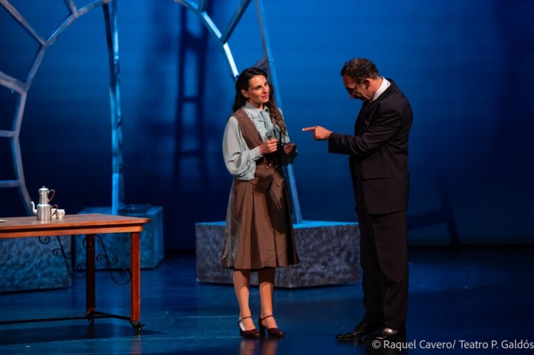 Dos intérpretes de pie junto a una mesa larga bajo luz azul; el le entrega algo a ella. Foto: Raquel Cavero / Teatro Pérez Galdós