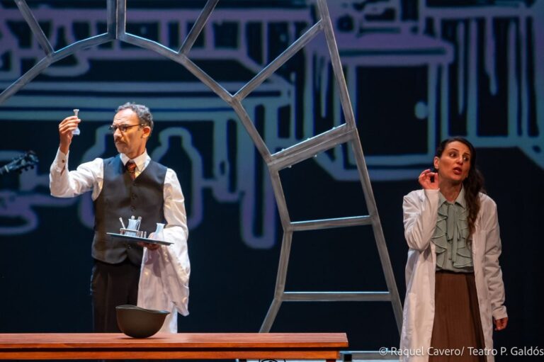 Un intérprete de bata blanca alza un vaso junto a una compañera, ante la escenografía de trazos. Foto: Raquel Cavero / Teatro Pérez Galdós