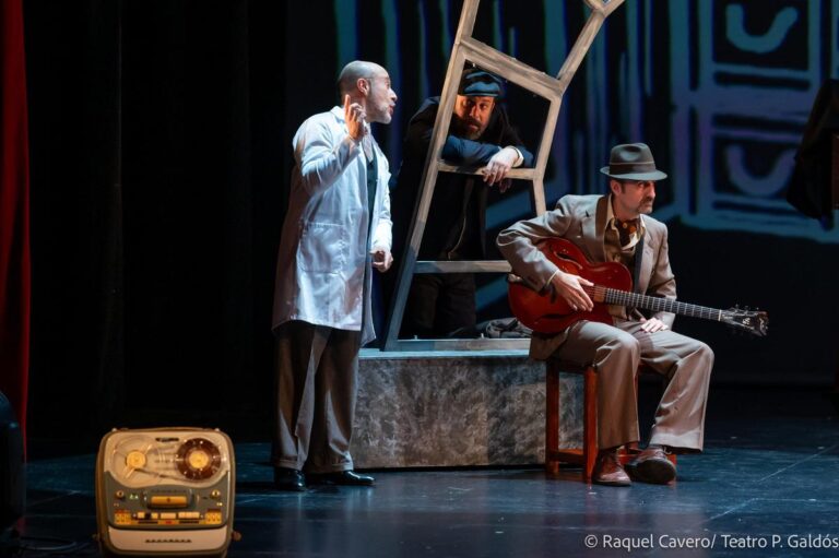 Un intérprete de bata blanca de pie junto al guitarrista sentado y un tercer personaje con sombrero. Foto: Raquel Cavero / Teatro Pérez Galdós