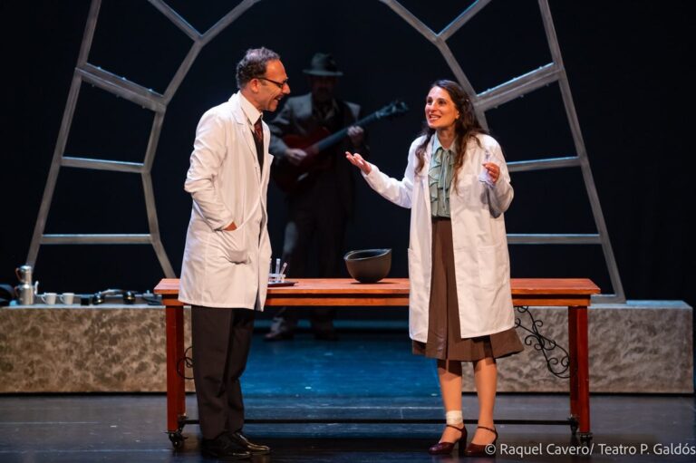 Dos intérpretes de bata blanca discuten a un lado y otro de la mesa del laboratorio. Foto: Raquel Cavero / Teatro Pérez Galdós