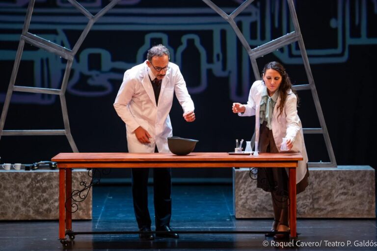 Dos intérpretes de bata blanca trabajando inclinados sobre la mesa del laboratorio. Foto: Raquel Cavero / Teatro Pérez Galdós