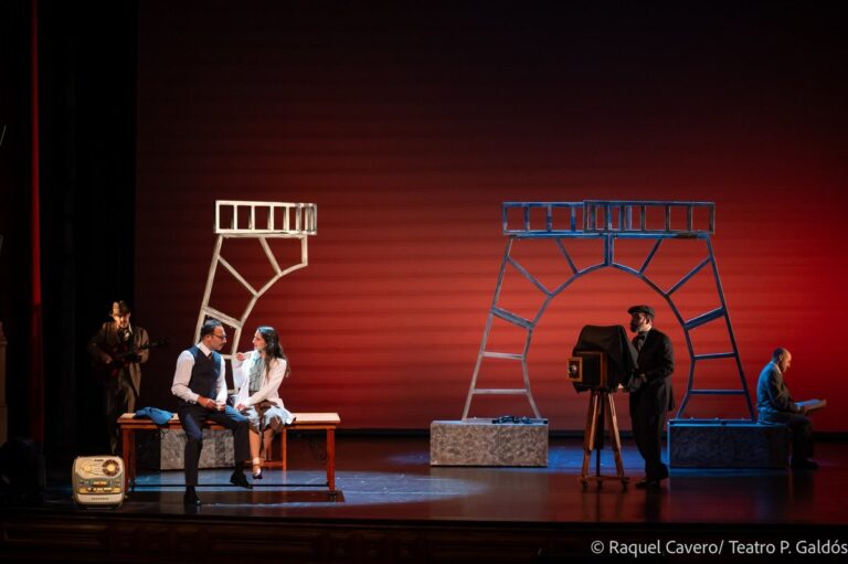 Escena sobre fondo rojo con la cámara de fuelle a un lado y varios personajes sentados. Foto: Raquel Cavero / Teatro Pérez Galdós