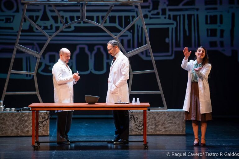 Tres intérpretes de bata blanca en torno a la mesa del laboratorio, uno con el brazo en alto. Foto: Raquel Cavero / Teatro Pérez Galdós