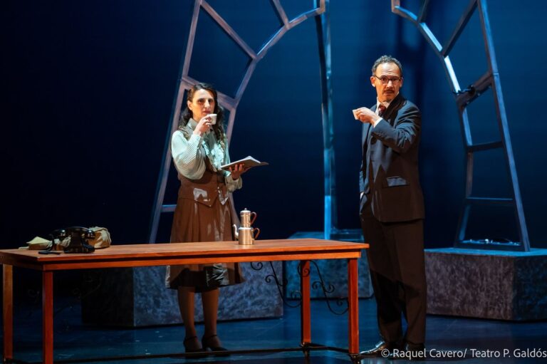 Una intérprete lee un libro mientras otro personaje la señala desde el otro lado de la mesa. Foto: Raquel Cavero / Teatro Pérez Galdós