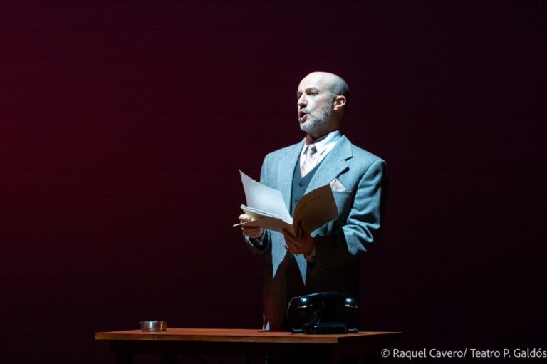 Un intérprete de traje claro lee un documento de pie sobre la mesa. Foto: Raquel Cavero / Teatro Pérez Galdós