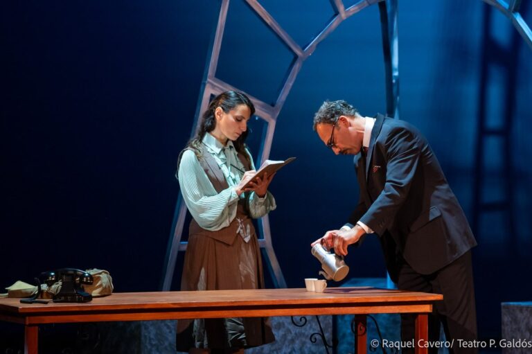 Un intérprete sirve algo en un vaso mientras una compañera lee un cuaderno al otro lado de la mesa. Foto: Raquel Cavero / Teatro Pérez Galdós