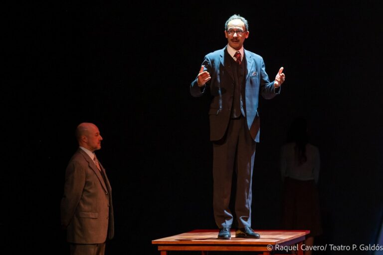Un intérprete de traje, de pie sobre la mesa, se dirige al público gesticulando. Foto: Raquel Cavero / Teatro Pérez Galdós