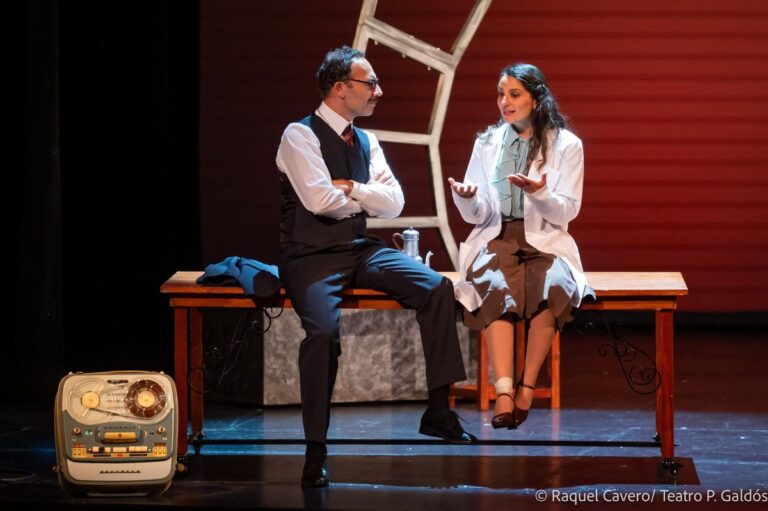 Dos intérpretes sentados en el borde de la mesa, ella con bata blanca, junto al magnetófono. Foto: Raquel Cavero / Teatro Pérez Galdós