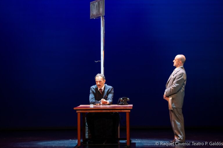 Un intérprete sentado al escritorio y otro de pie frente a el, sobre fondo azul. Foto: Raquel Cavero / Teatro Pérez Galdós